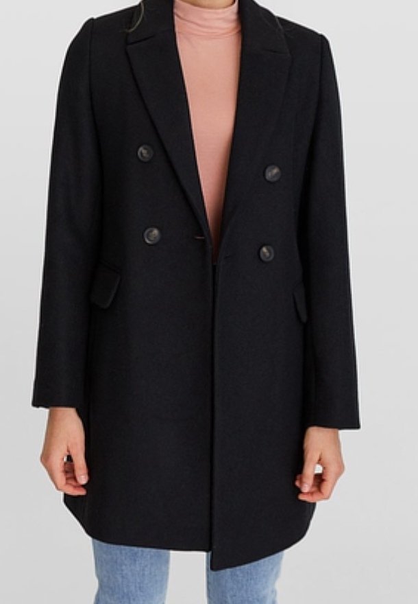 Femme portant un manteau noir croisé sur un col roulé rose et un jean bleu clair, debout devant un fond uni.