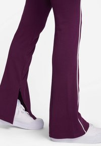 Pantalons violets évasés avec des zips latéraux et des rayures blanches contrastantes, associés à des baskets blanches. Tissu doux avec une texture lisse.