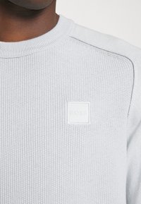 Jasnoszary sweter dziewiarski z prążkowanym kołnierzem, z kwadratową białą łatką z logo. Teksturowana tkanina z delikatnymi detalami ściegów.