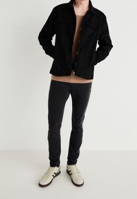 Musta mokkanahkainen takki, jossa on kaksi rintataskua, vaaleanbeige paita alla, tummat farkut ja vaaleat tennarit tummilla yksityiskohdilla. Yleinen pinta on sileä.