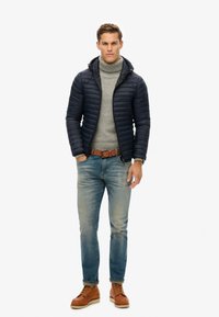 Marineblå dunjakke med hætte, grå turtleneck, blå jeans og brune snørebåndsstøvler. Har diskrete syninger og en afslappet pasform.