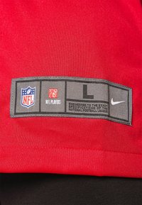 Un tejido rojo con un acabado texturizado presenta una etiqueta gris que muestra los logotipos de la NFL, 'Jugadores de la NFL', tamaño 'L' y la marca 'Nike'.