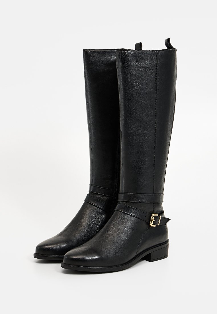 Schwarze knielange Lederstiefel mit flachen Absätzen und dekorativen Knöchelriemen mit goldfarbenen Schnallen, auf einem weißen Hintergrund präsentiert.