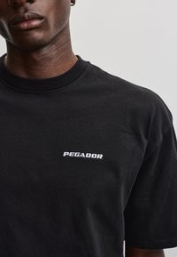 Detail osoby nosiacej čierne tričko s bielym nápisom „PEGADOR“ na hrudi a malým zlatým kruhovým náušnicou v ľavom uchu.