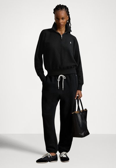 Juodas megztinis su užtrauktuku ir aukštu apykakliu, derinamas su juodomis sportinėmis kelnėmis su raišteliais. Juoda rankinė ir juodi batai su baltais raišteliais.