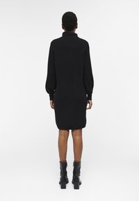 Schwarzes gestricktes Kleid mit hohem Kragen, langen Ärmeln und gerippter Textur, das knapp über dem Knie endet, kombiniert mit schwarzen Stiefeln mit Absatz.