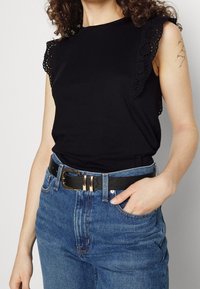 Sort ærmeløs top med blondedetaljer på skuldrene, parret med højtaljede blå denimjeans og et sort bælte med gyldne detaljer.