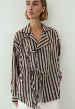 Femme aux cheveux blonds courts portant une chemise ample à rayures verticales marron et beige avec des liens sur le devant, debout devant un fond uni.