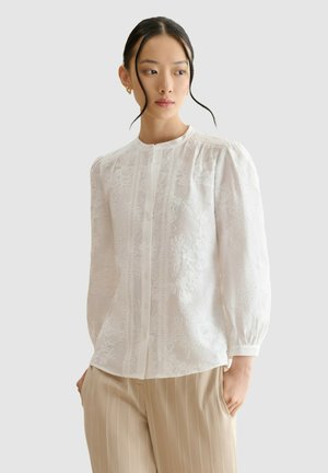 Mujer con una blusa blanca de manga larga con bordado floral y pantalones beige a rayas, de pie con una mano en el bolsillo frente a un fondo liso.