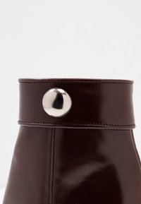 Botín de cuero marrón oscuro con puntera redondeada y un acabado brillante. Presenta un botón metálico plateado en la parte superior. Detalle de costura limpia.