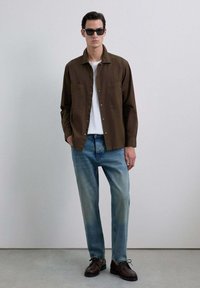 Bărbat purtând ochelari de soare negri, cămașă maro cu butoni peste un tricou alb, blugi și pantofi maro închis, stând în fața unui fundal simplu.