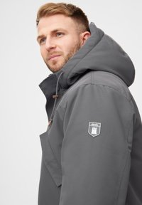 Uomo con capelli biondi che indossa una giacca grigia con cappuccio e un patch con il logo "derbe" sulla manica, che guarda leggermente verso l'alto e a sinistra.