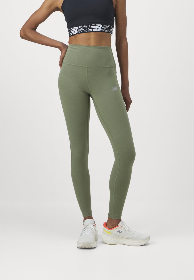 Leggings de talle alto en verde oliva con una textura suave, que presentan un sutil logotipo en el muslo y se combinan con zapatillas deportivas blancas.