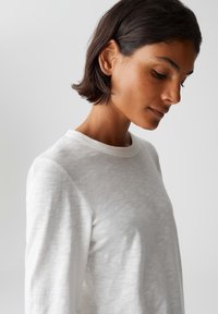 Donna con i capelli corti e scuri guarda in basso, indossando una semplice maglietta bianca a maniche lunghe su uno sfondo grigio chiaro uniforme.