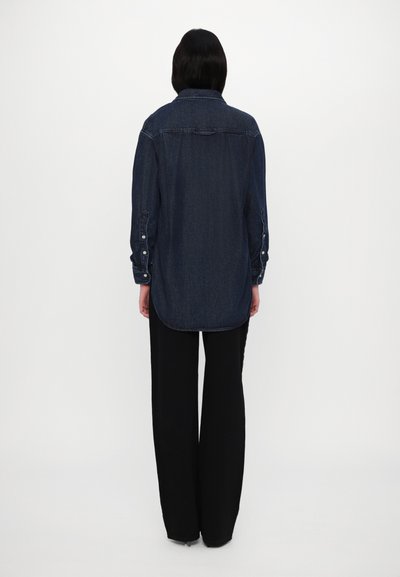 Chemise en denim bleu foncé, avec une fermeture à boutons sur le devant, des manches longues et un ourlet incurvé, associée à un pantalon large noir.