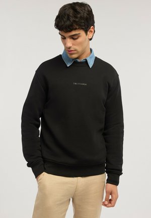 Junger Mann trägt schwarzen Trussardi-Pullover über blauem Hemd mit Kragen, beige Hose, steht mit teilweise in den Taschen steckenden Händen und schaut nach unten.