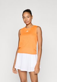 Πορτοκαλί αμάνικη αθλητική μπλούζα με κάθετες ρίγες, σε συνδυασμό με λευκή φούστα. Η μπλούζα έχει στρογγυλή λαιμόκοψη και διακριτικό λογότυπο.