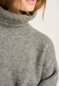 Pull en col roulé gris en tissu tricoté texturé avec des côtes verticales. La vue en gros plan met en avant le col et la finition lisse.