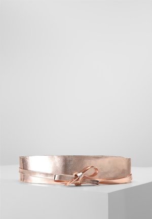 Roze-gouden metalen riem met smalle riem vastgeknoopt in een strik, tentoongesteld op een wit voetstuk tegen een effen lichtgrijze achtergrond.