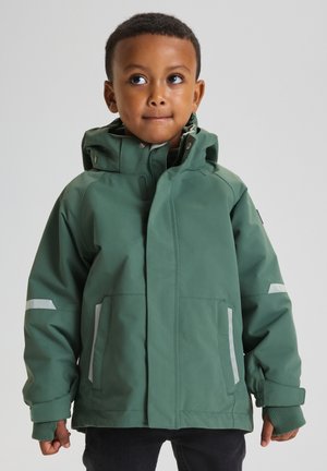 Jong kind met een groen waterdicht jack met capuchon, staand en omhoog kijkend naar de zijkant tegen een effen achtergrond.