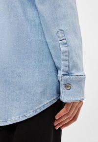 Ανοιχτό μπλε τζιν πουκάμισο με κουμπωμένα μανίκια, που διαθέτει ασημένιο κουμπί, ορατά ραφές και ίσια στρίφωμα. Μαλακή υφή και χαλαρός σχεδιασμός.