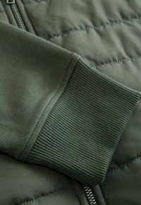 Grüner gesteppter Jackenstoff mit gerippten Manschetten. Das Material ist weich und zeigt eine glatte Textur sowie gleichmäßige Nähte entlang der Nähte.