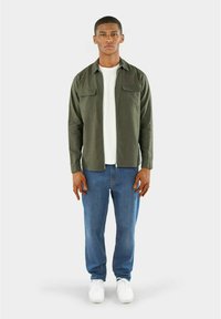 Donker groene, knoopsluiting overhemd met twee borstzakken, gecombineerd met een witte T-shirt en lichtblauwe jeans. Witte sneakers maken de outfit compleet.