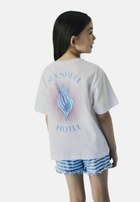 Witte katoenen t-shirt met een blauwe schelpgrafiek en de tekst "SEA SHELL HOTEL", gecombineerd met blauw-wit gestreepte rucheshort.