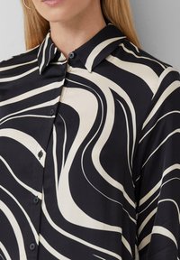 Donna che indossa una camicia button-up con un motivo astratto a spirale bianco e nero, con colletto e orecchino a cerchio.