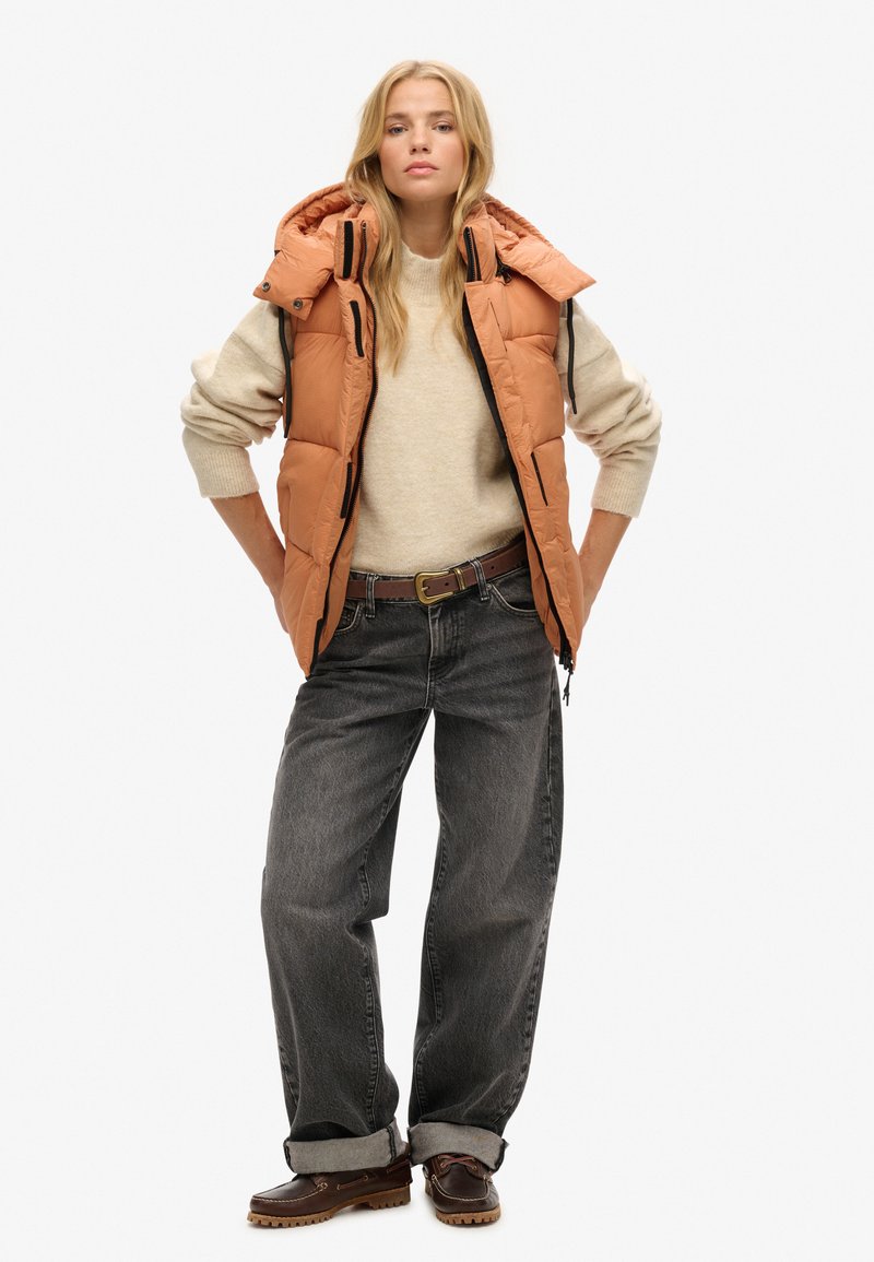 Kamizelka puchowa w ciepłym pomarańczowym kolorze z kapturem, zestawiona z beżowym swetrem i luźnymi szarymi dżinsami. Brązowe obuwie z teksturowaną podeszwą.