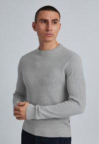 Bărbat cu păr scurt, purtând un pulover tricotat cu textură de culoare gri deschis, cu mâinile strânse, privind în lateral pe un fundal gri simplu.