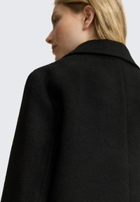 Cappotto di lana nero con una finitura testurizzata, dotato di collettore a punto e spalle strutturate. La schiena è parzialmente visibile, evidenziando linee pulite.
