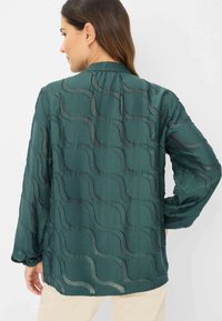 Dark green, sheer blouse with wavy texture pattern, long sleeves, and a mandarin collar, featuring a relaxed fit and subtle sheen.