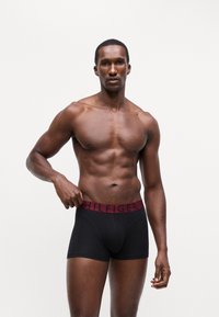 Črne bokserice z bordo elastičnim pasom, na katerem je v črni barvi napisan "HILFIGER". Model ima atletsko postavo in je brez majice.