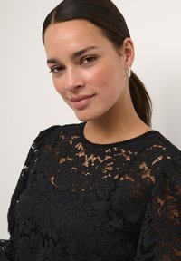 Must mustriga tops, millel on lillemustrid, läbinägelik tekstuur ja ümmargune dekoltee. Modellil on pikad juuksed tagasi seotud ja ta kannab rõngakujulisi kõrvarõngaid.