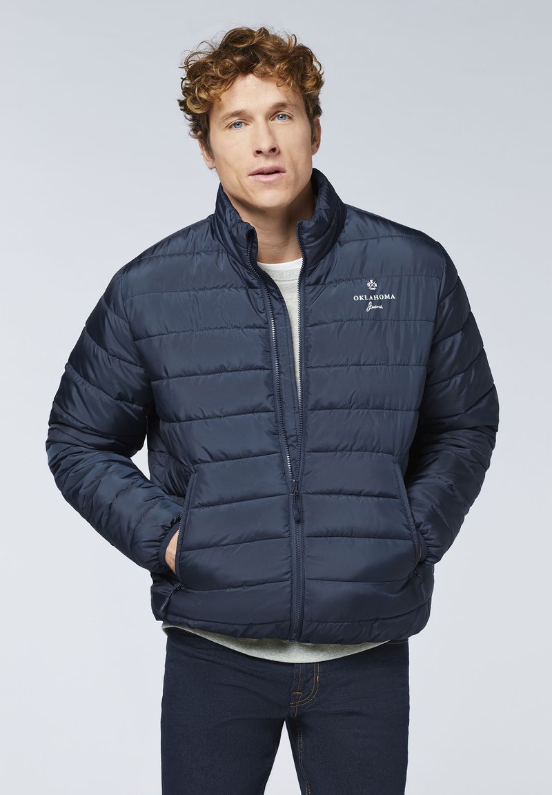 Marineblaues Steppjäckchen mit Reißverschluss, gestepptem Design und seitlichen Taschen. Verfügt über ein gesticktes Logo auf der linken Brust.