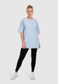 Camiseta oversized de color azul claro con mangas cortas, de material de algodón, que presenta un pequeño logo blanco. Combinada con leggings negros y zapatillas blancas.