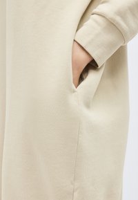 Χέρι στην πλαϊνή τσέπη ενός μπεζ ενδύματος με μακριά μανίκια από απαλό, ριγωτό ύφασμα.