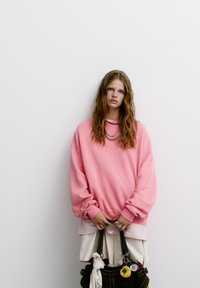 Rózsaszín, oversized kapucnis pulóver, bordás mandzsettával, fehér inggel rétegezve, barna táskát tartva, amelyen különböző kulcstartók és gyűrűk találhatók.