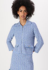 Helehelesin sinine tweed jakk, millel on tõmblukk, krae ja kaks esimest taskut. Tekstuuriga kangas koos kootud mustrite ja õrna sädelusega.
