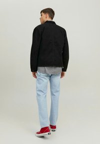 Giacca in suede nera con colletto, indossata sopra una camicia a quadri, abbinata a jeans azzurri e sneaker rosse. Vista posteriore, texture liscia.