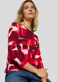 Frau mit blondem Haar, die ein langärmliges Oberteil mit rotem und weißem abstraktem Muster und blaue Jeans trägt, posiert vor einem einfarbigen Hintergrund.