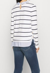 T-shirt à rayures à manches longues en blanc et bleu marine, avec un col bleu clair, des fentes latérales ornées de boutons décoratifs et un détail en forme de trou de clé au dos.