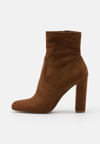 Botín de ante marrón hasta el tobillo con un tacón alto y bloque y punta redondeada, con costuras visibles y cierre de cremallera lateral.