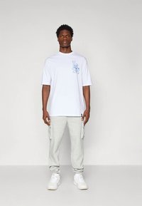 Baltas marškinėlis su mėlynu grafiniu logotipu krūtinėje, derinamas su šviesiai pilkomis sportinėmis kelnėmis su šoninėmis kišenėmis ir baltais sportiniais batais.