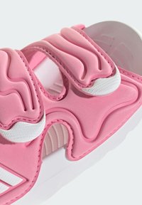 Różowa sandał z falistym, teksturowanym wierzchem, z białymi akcentami i paskami na rzepy. Podeszwa jest biała o gładkim wykończeniu.