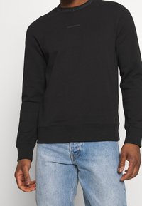 Sweat-shirt noir en coton avec un col rond, orné d'un logo imprimé au niveau du col et des manches longues. Associé à un jean bleu clair.