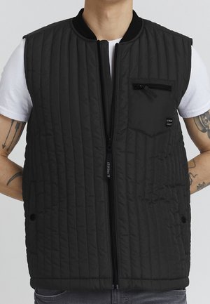 Veste matelassée noire avec fermeture éclair frontale et poche zippée sur la poitrine portée par-dessus une chemise blanche à manches courtes par une personne ayant des tatouages sur les deux bras.