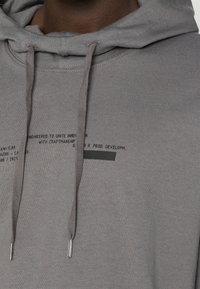 Sudadera gris de tela suave, con capucha ajustable, puños acanalados y una etiqueta impresa en el pecho con texto negro.