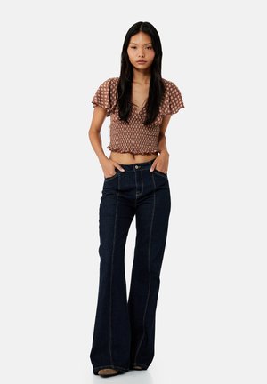 Frau mit langen schwarzen Haaren, die ein braunes gemustertes kurzes Oberteil und dunkle, hochgeschnittene Schlagjeans trägt, Hände in den Taschen, neutrale Mimik.